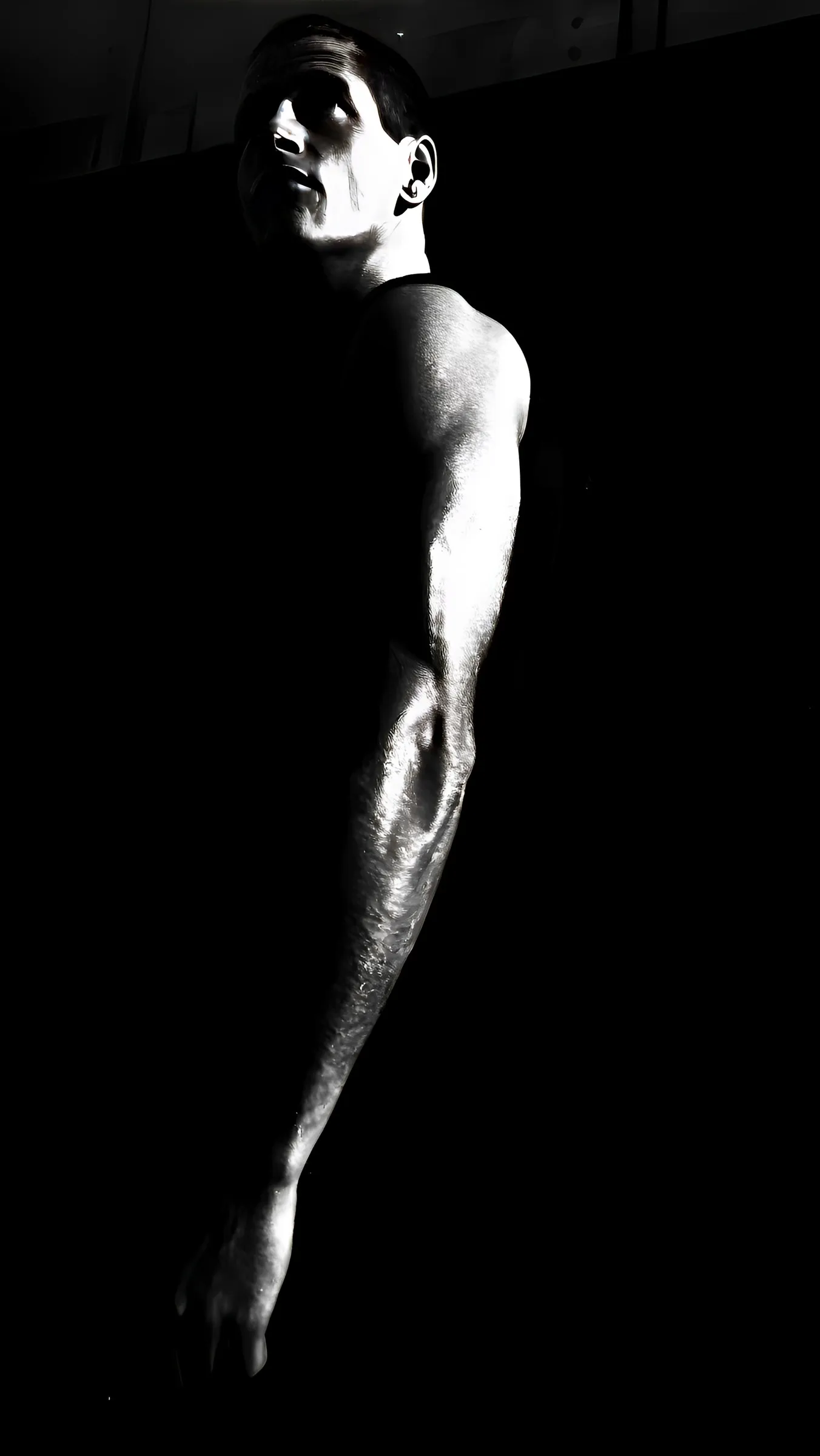 Black-and-white photograph of a shirtless man seen in profile from behind, his head tilted back, with dramatic side lighting illuminating his face, shoulder, and extended arm against a black background.