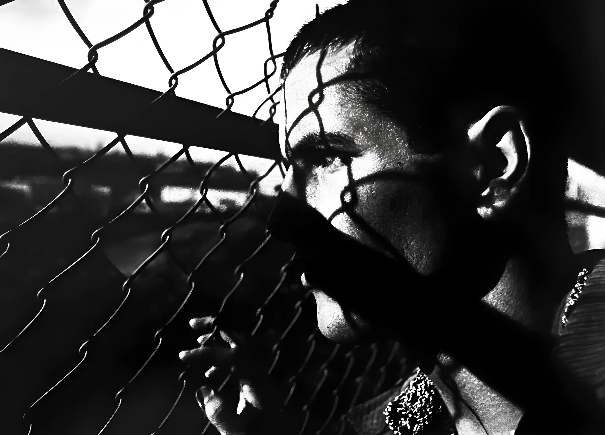 Black-and-white close-up portrait of a person's face pressed close to a chain-link fence, one hand raised near the fence, with strong directional light and shadow across the face.