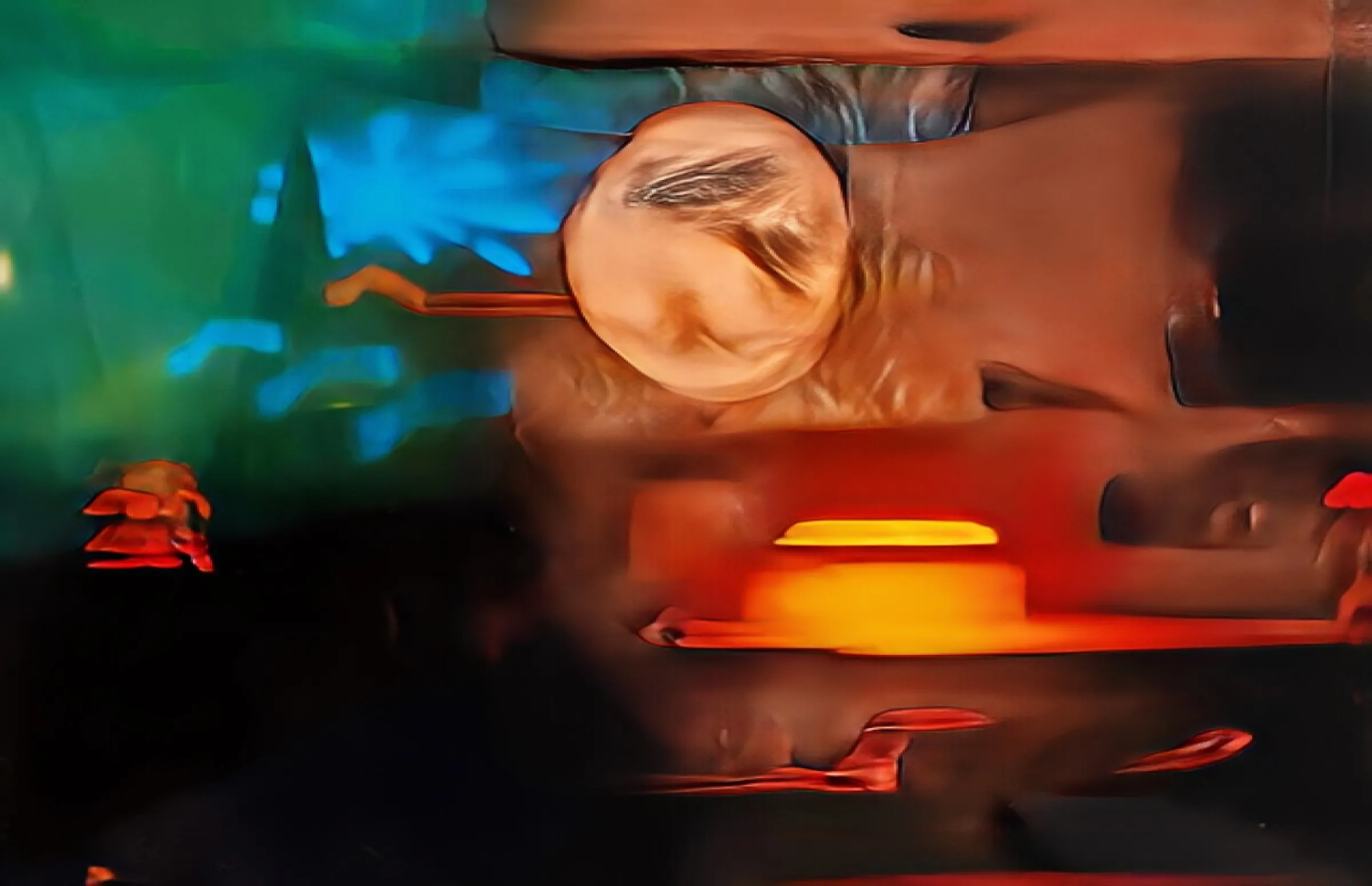 Abstract, blurred long-exposure color photograph with swirling green, blue, orange, and yellow tones and a glowing circular highlight at center, suggesting an interior with warm and cool light sources.