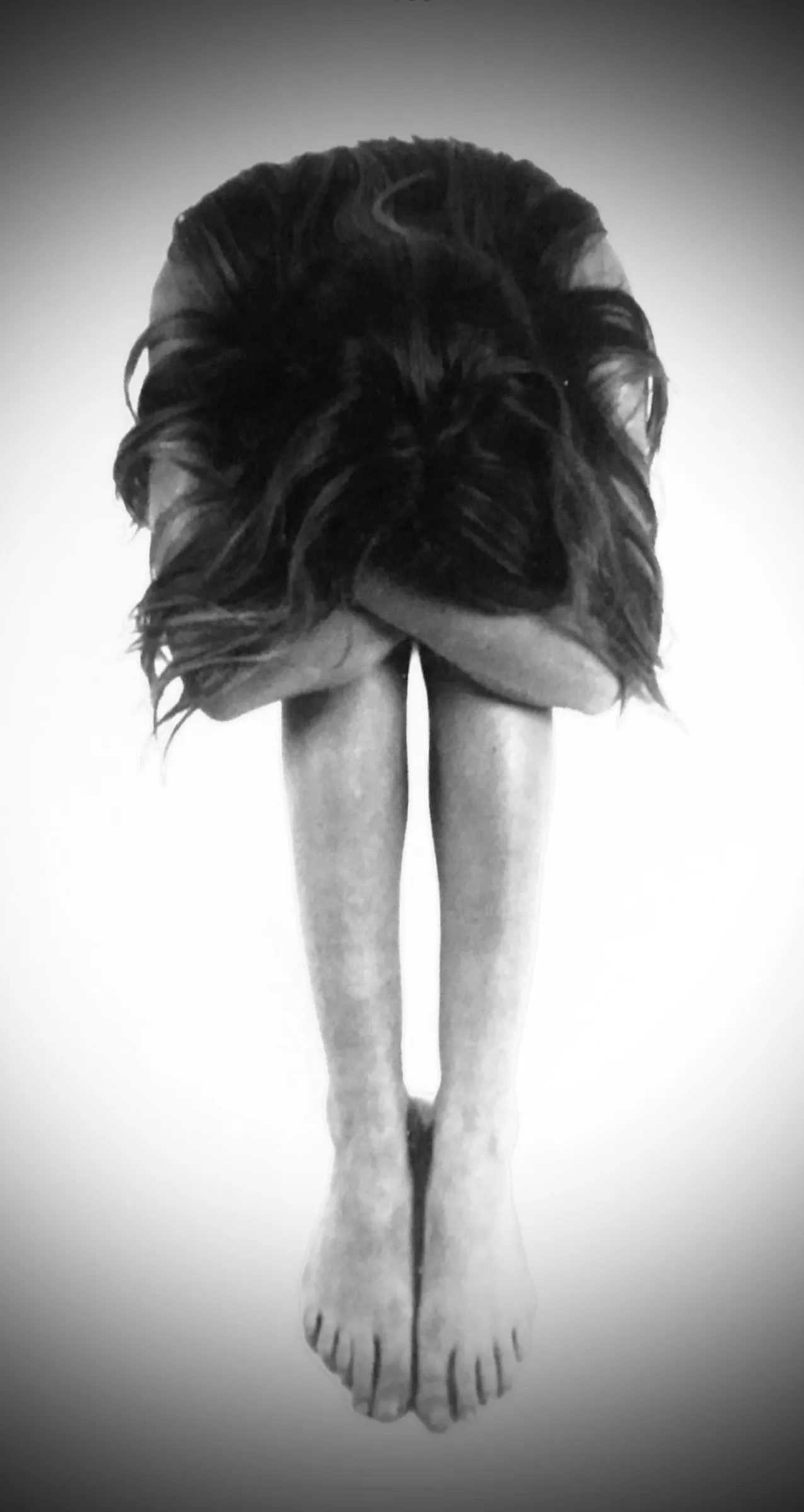 Black-and-white photograph of a person bent forward with long dark hair covering their face, arms wrapped around their knees, bare legs and feet visible, set against a soft white vignette background.