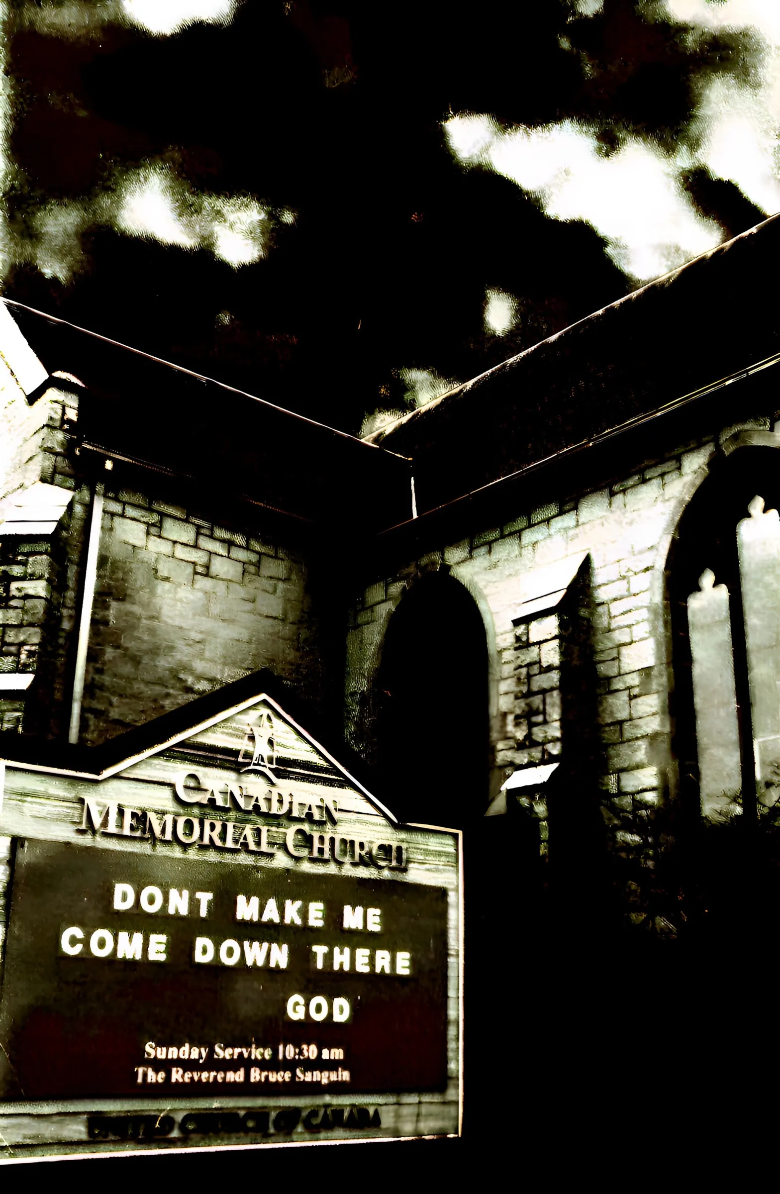 Black-and-white photograph of a stone church at night under a dramatic cloudy sky, with an illuminated sign reading "Canadian Memorial Church, Don't Make Me Come Down There, God, Sunday Service 10:30 am, The Reverend Bruce Sanguin."