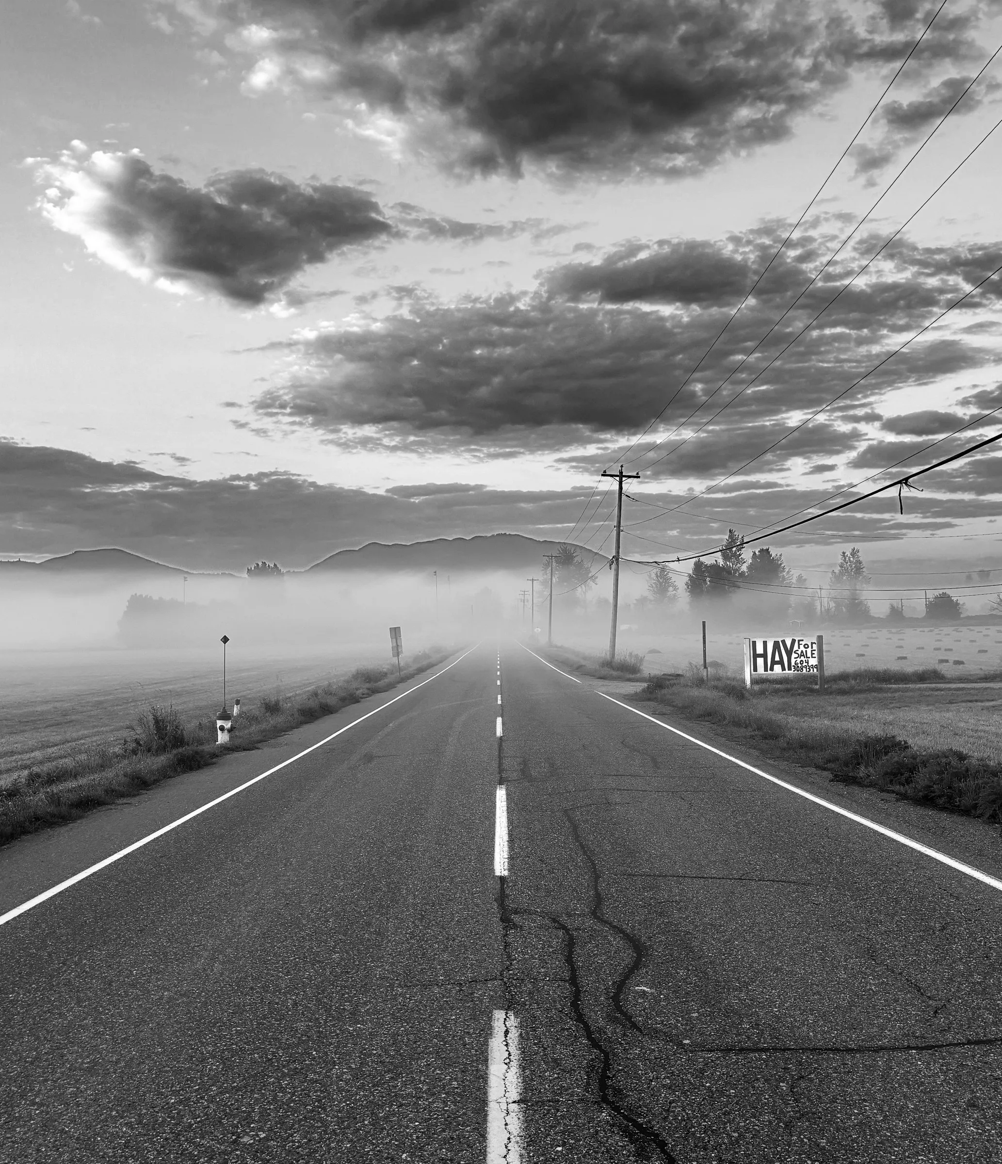 Black-and-white photograph of a foggy rural road stretching toward misty hills, with power lines, utility poles, and a roadside sign reading "HAY" visible along the shoulder.