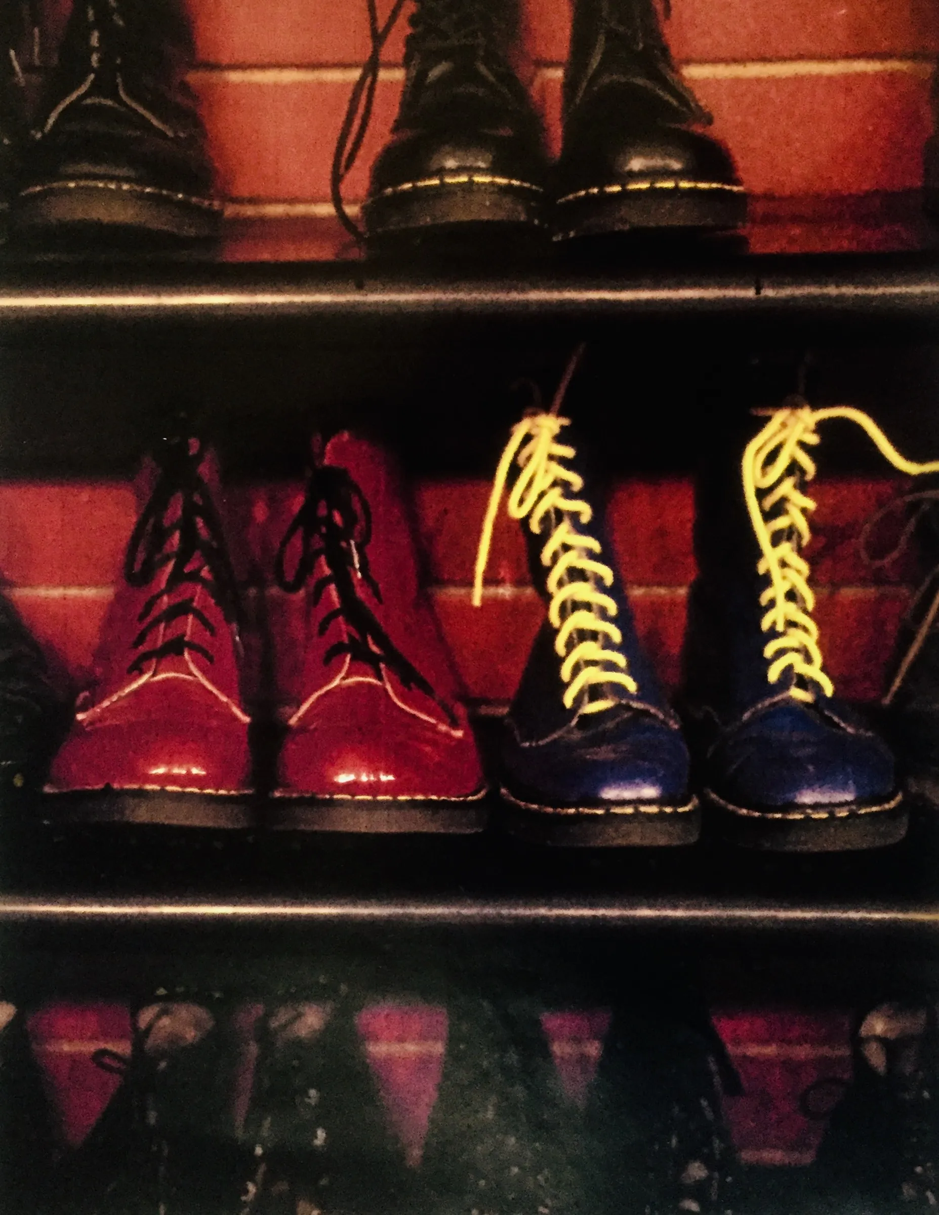 Color photograph looking down at three pairs of lace-up leather boots -- black, deep red with black laces, and navy blue with yellow laces -- lined up on wooden shelves against a red-striped wall.