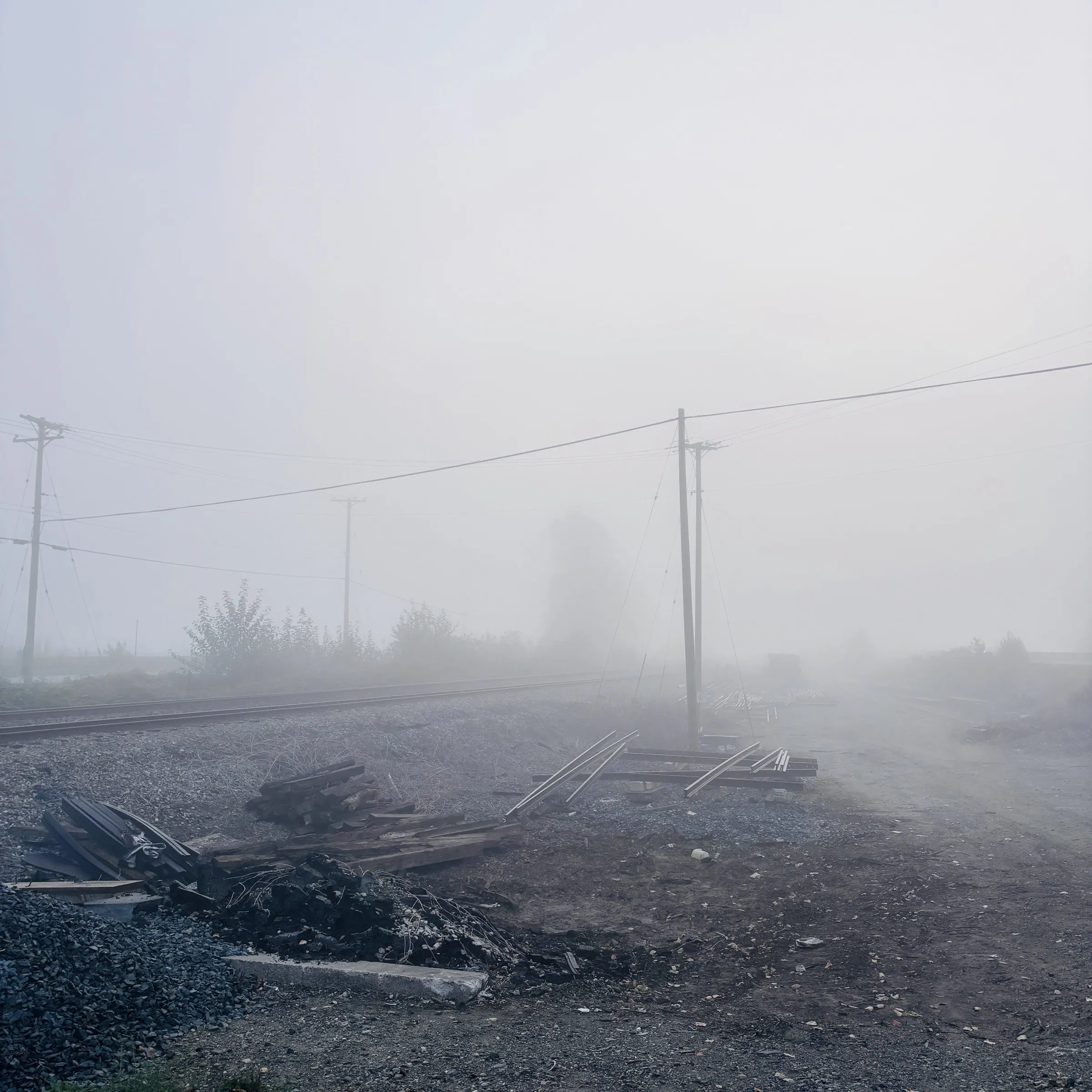 Photograph of a foggy industrial railway yard at dawn, with silhouetted utility poles, scattered rail ties and debris on the ground, and a hazy tree fading into the mist.