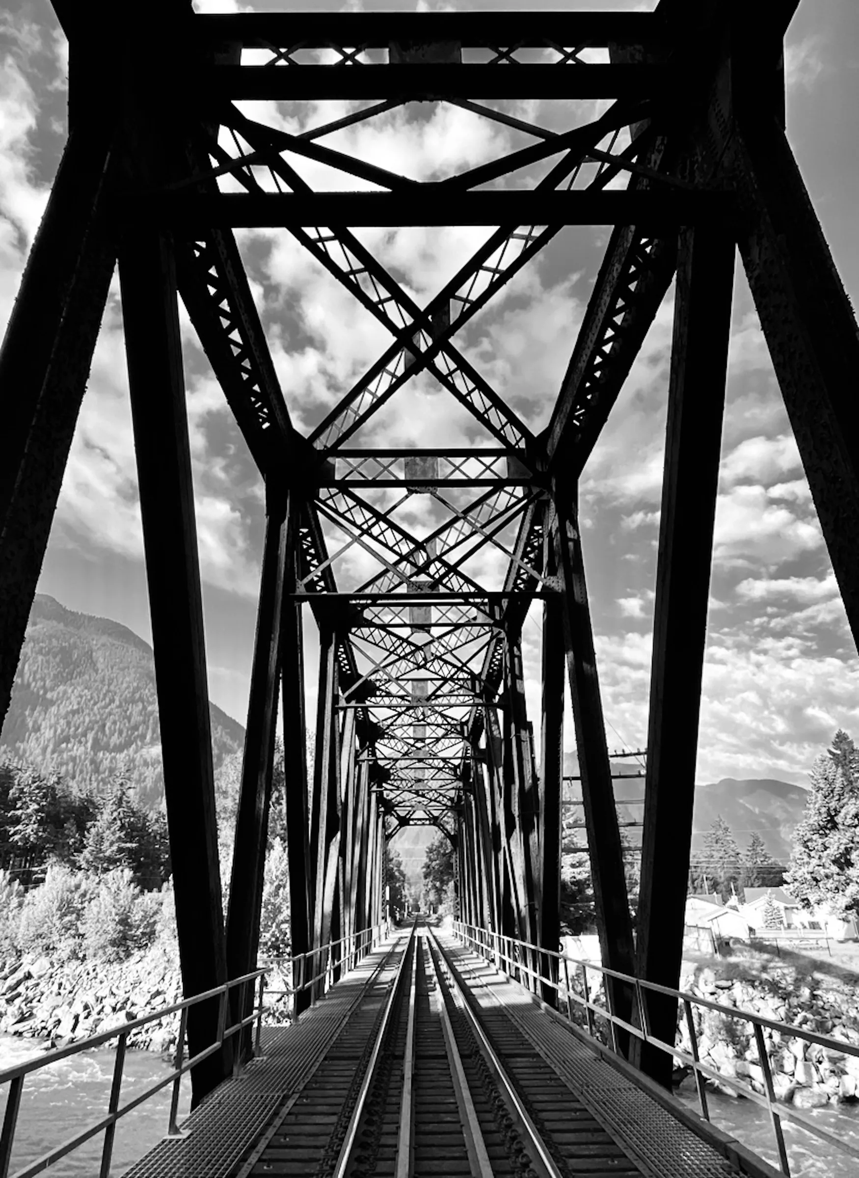 Black-and-white photograph looking down the length of a steel truss railway bridge toward mountains and clouds, with the railway tracks receding into the center of the frame.