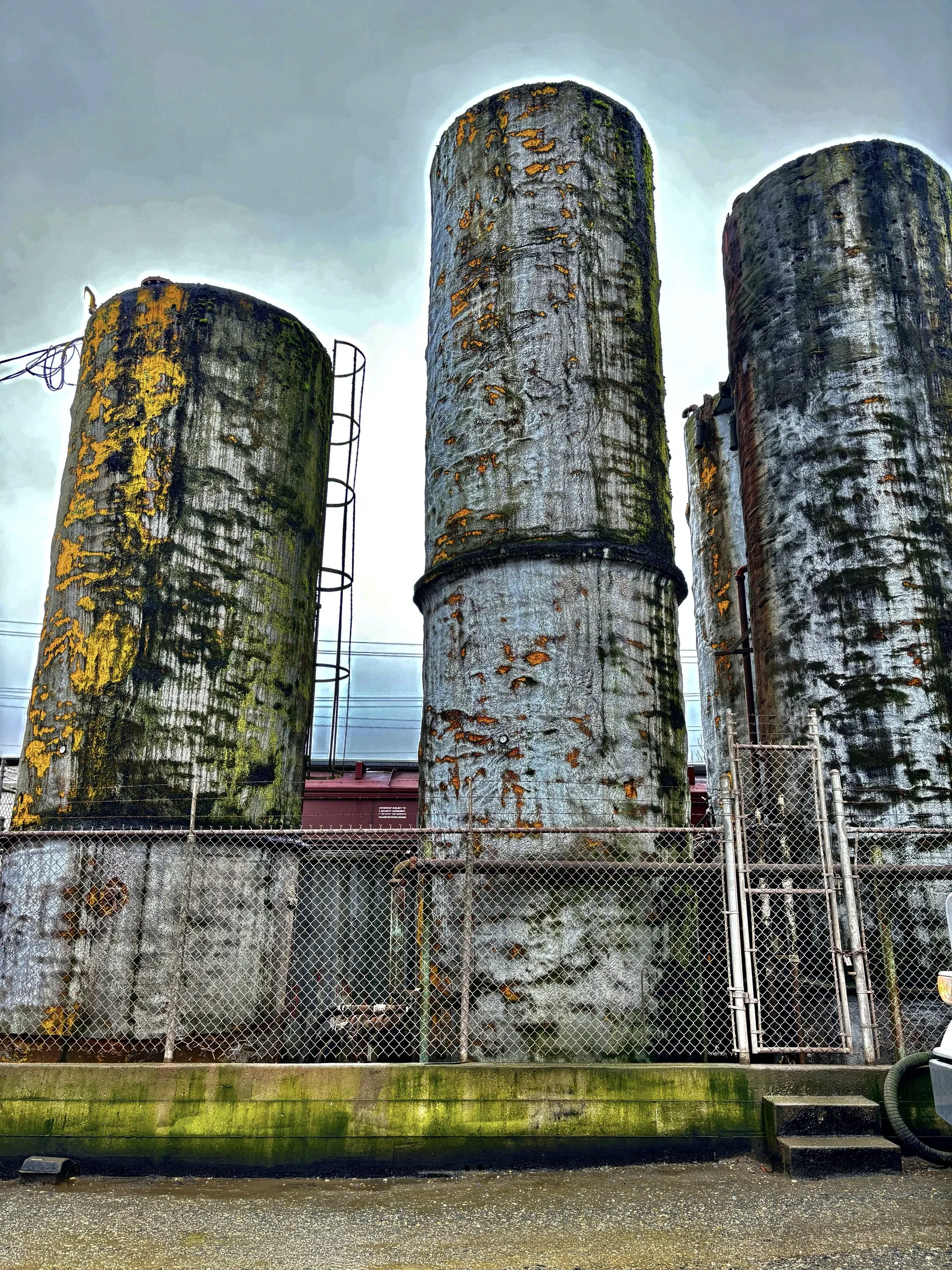 Color photograph of three tall, weathered concrete silos streaked with rust and lichen, standing behind a chain-link fence with a red rail car visible in the background.