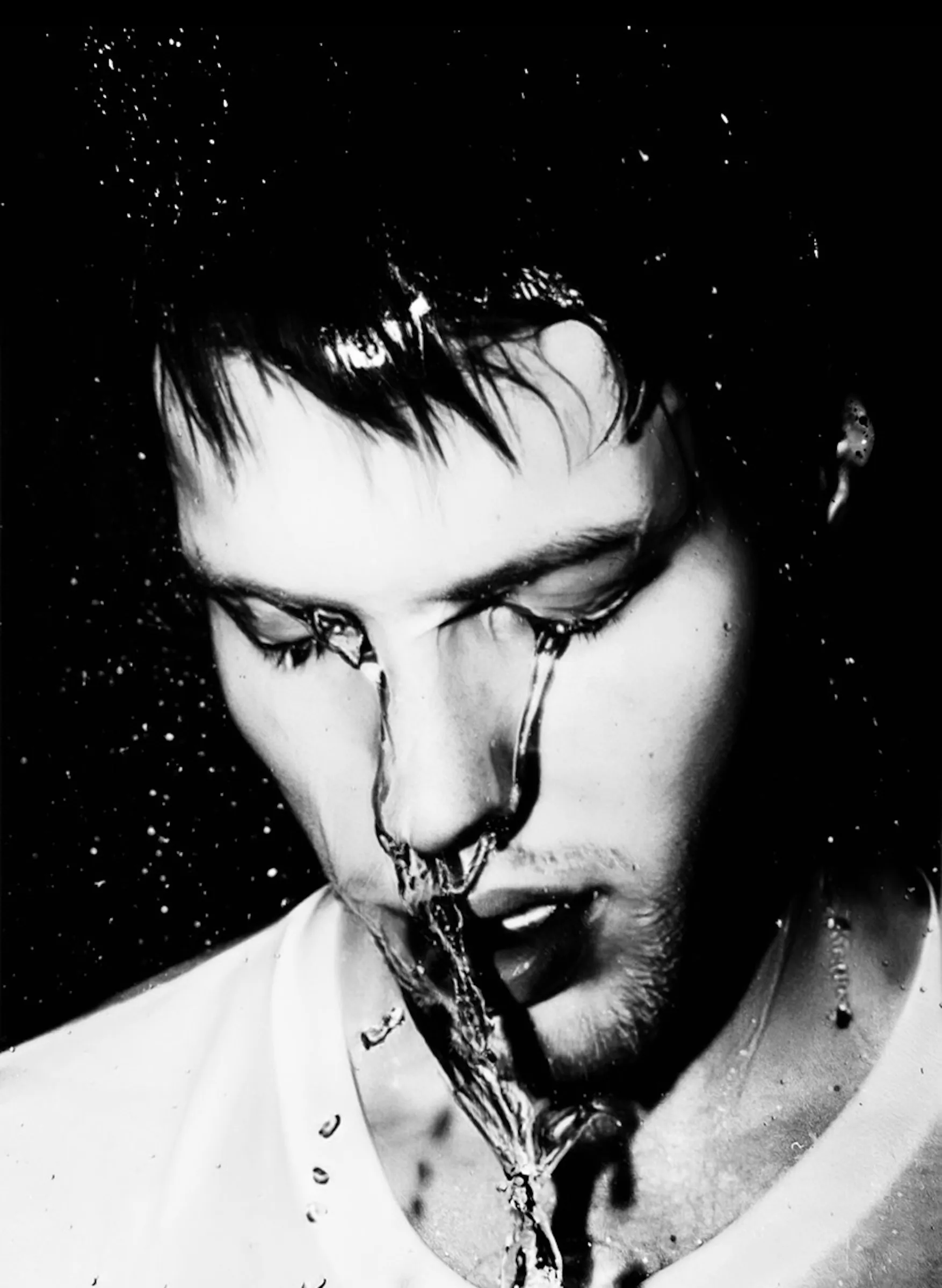 Black-and-white close-up portrait of a young man with wet hair and face, water streaming down his forehead, nose, and chin onto his white shirt, against a dark background.
