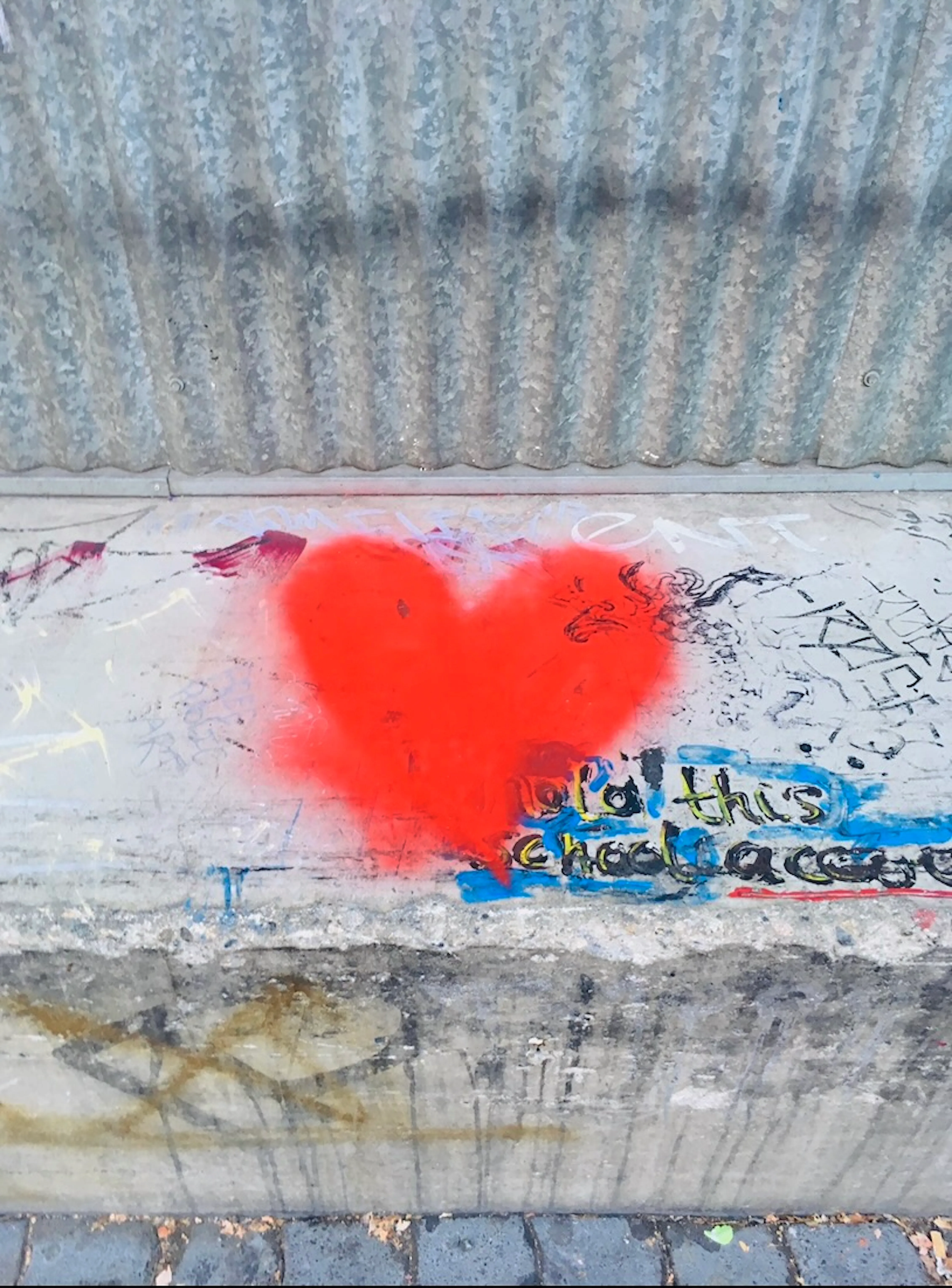 Weathered corrugated metal wall above a concrete curb covered in graffiti, with a red spray-painted heart above partially legible painted text.