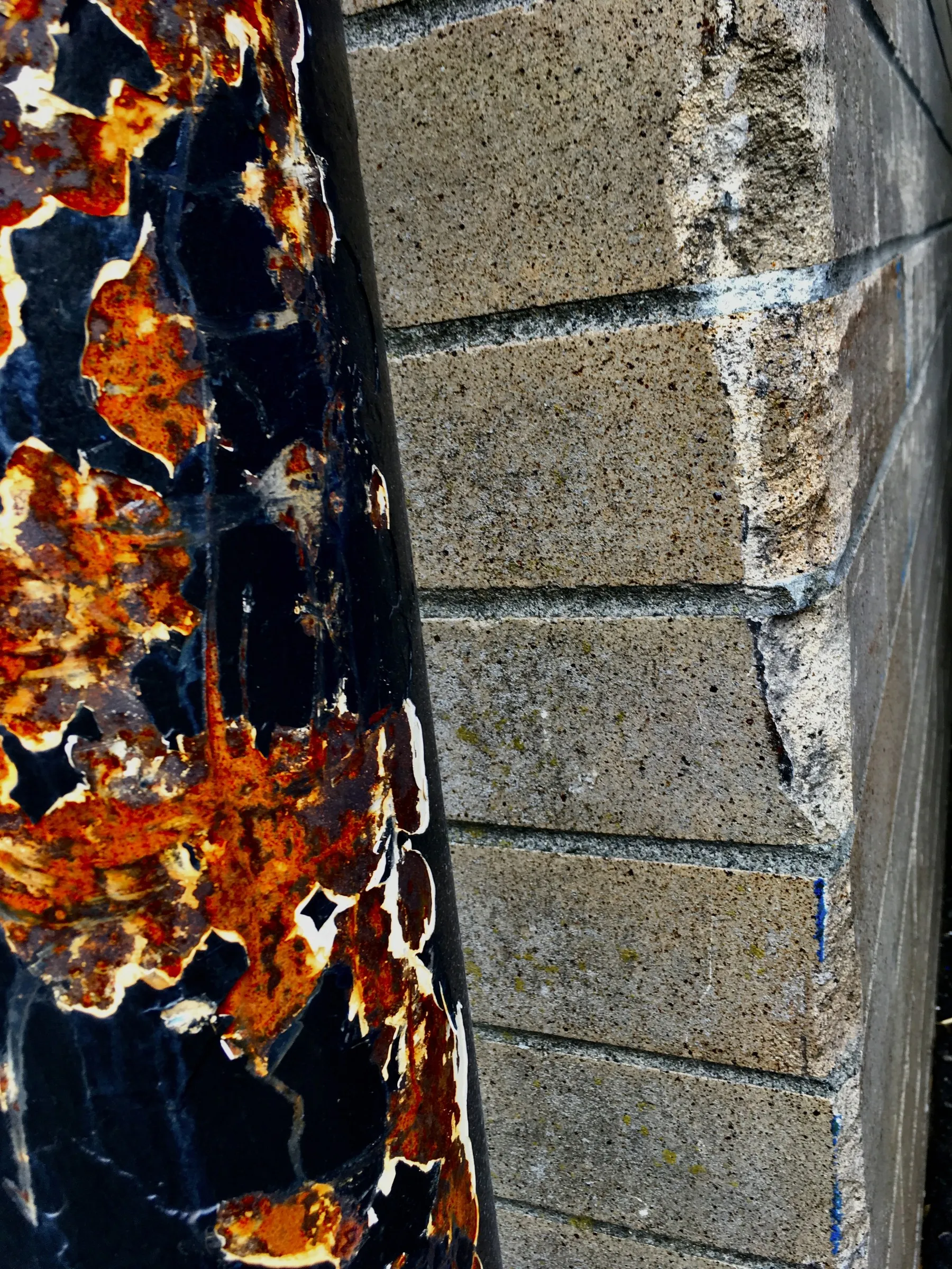 Close-up color photograph of a weathered rusted metal surface with peeling black paint and orange rust patches beside a textured tan stone wall.