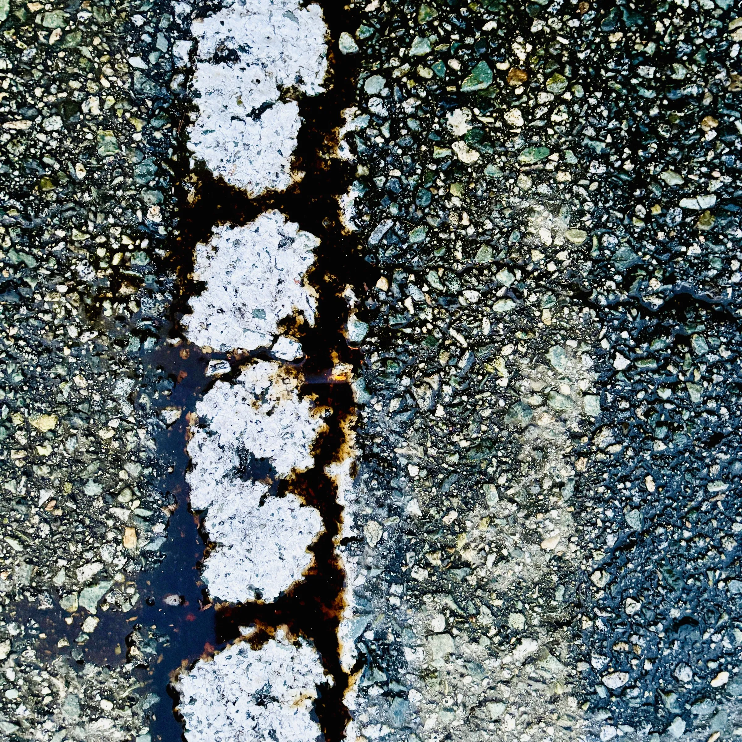 Overhead color photograph of cracked pavement with light-colored patching compound running through a dark seam, surrounded by speckled gravel and asphalt.