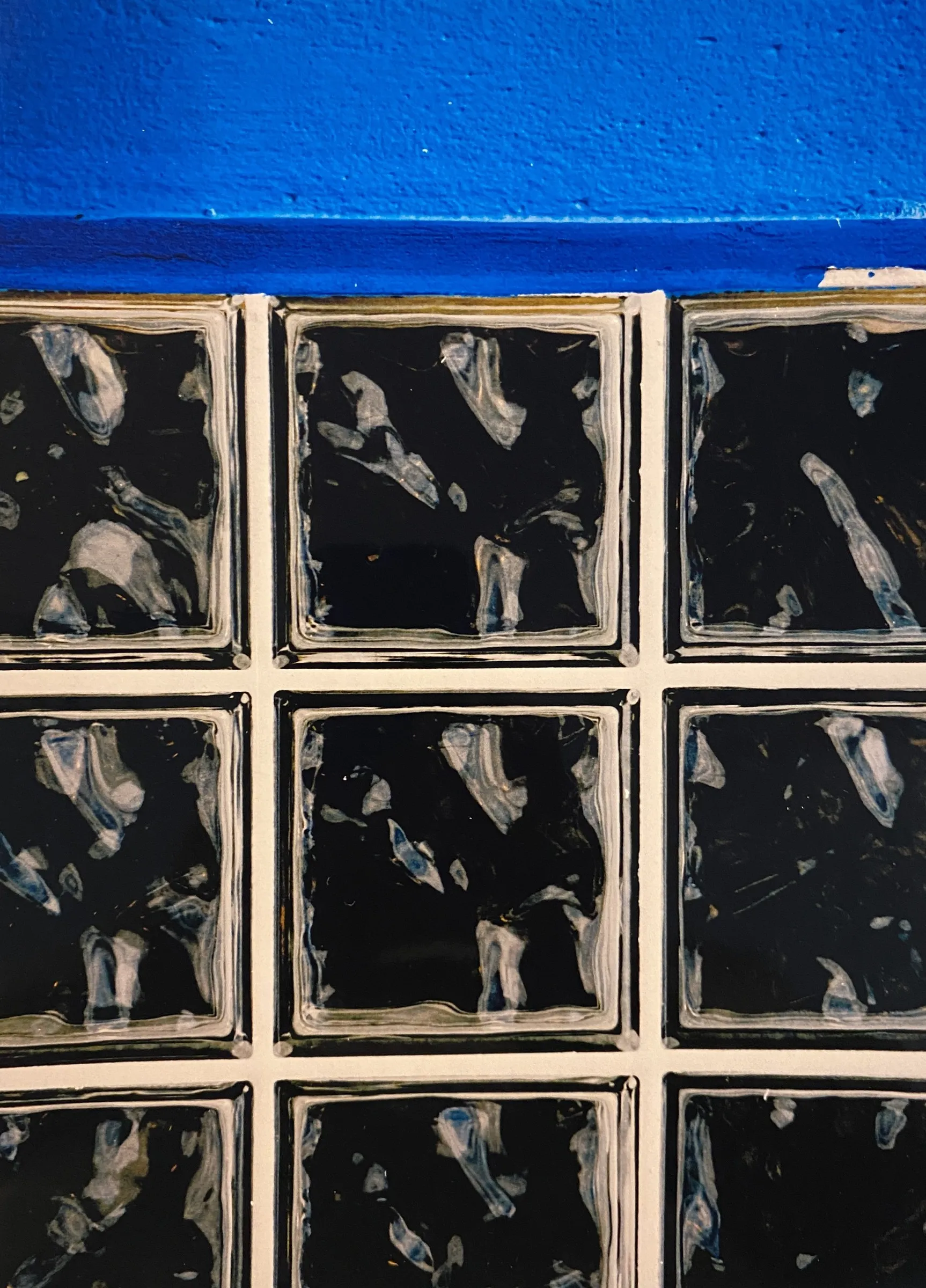 Close-up color photograph of a wall of glass blocks with mottled dark patterns, topped by a band of glossy blue paint.