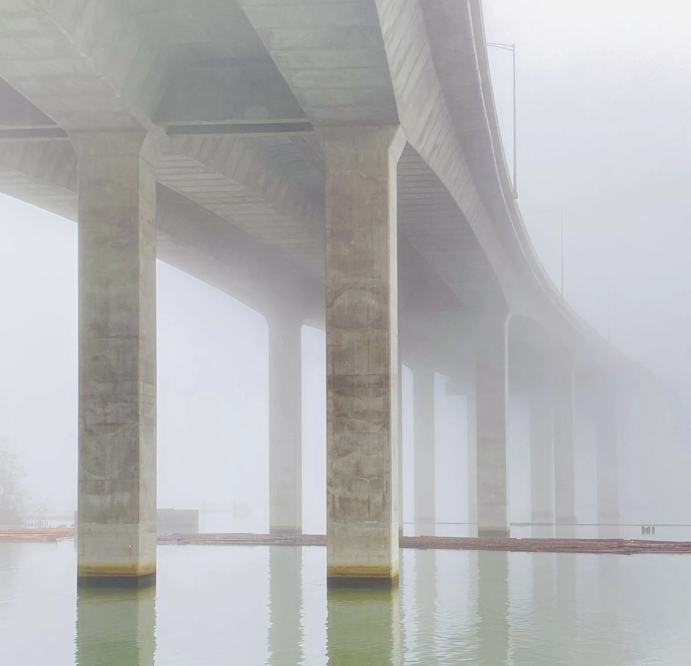 Color photograph of a concrete highway overpass and its support columns disappearing into thick fog above a calm body of water.