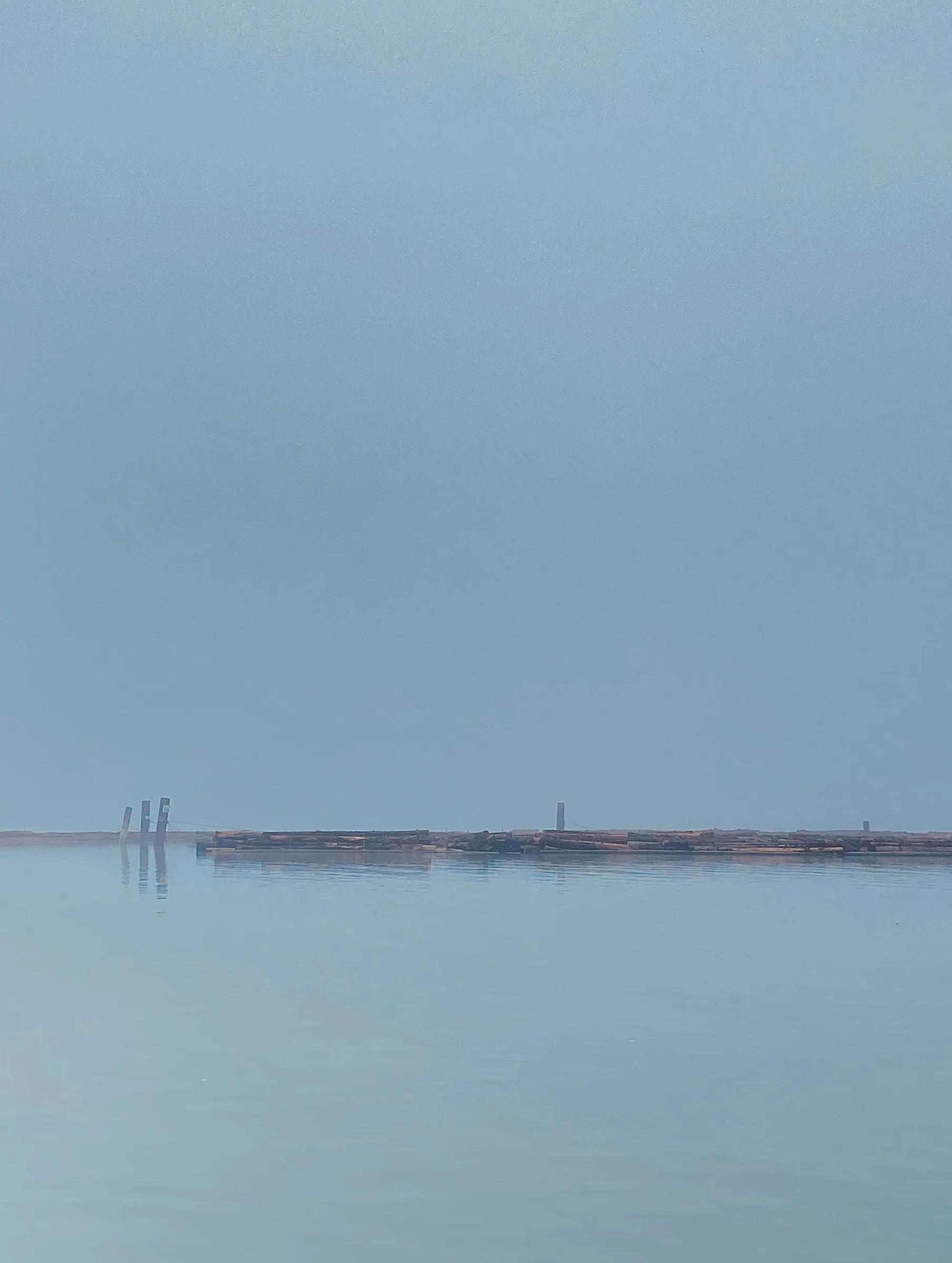 Minimalist color photograph of a low wooden dock or log boom barely visible on a foggy horizon line between pale sky and calm water.