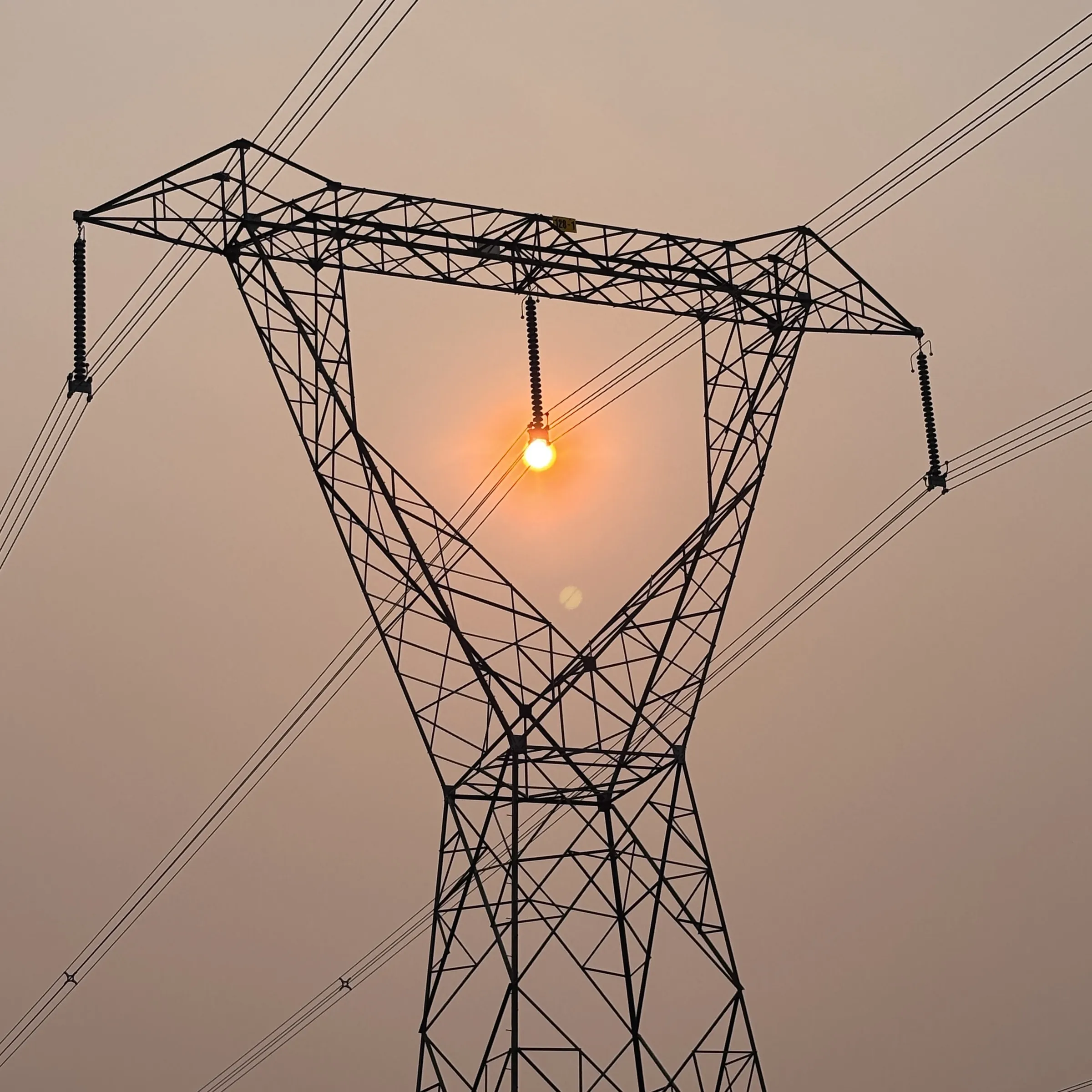 Silhouette photograph of an electrical transmission tower against a warm, hazy sky, with the setting sun glowing directly behind one of its insulator strings.