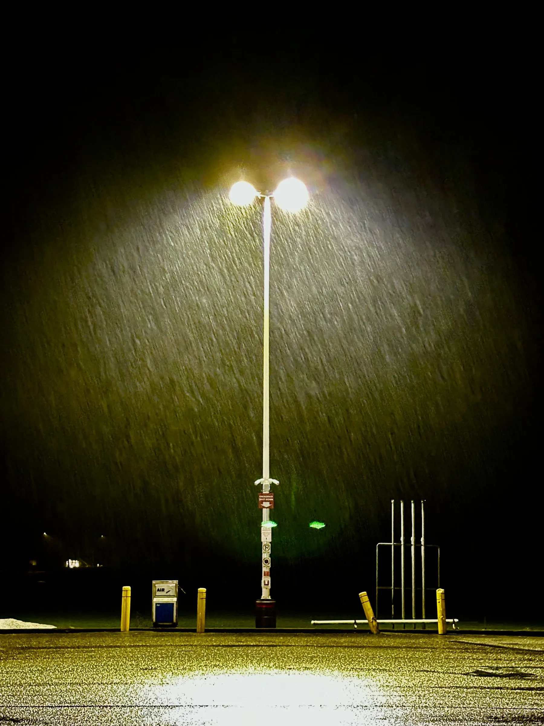 Color night photograph of a tall parking-lot light pole illuminating falling rain or snow, with bollards and a metal structure visible in the dark lot below.
