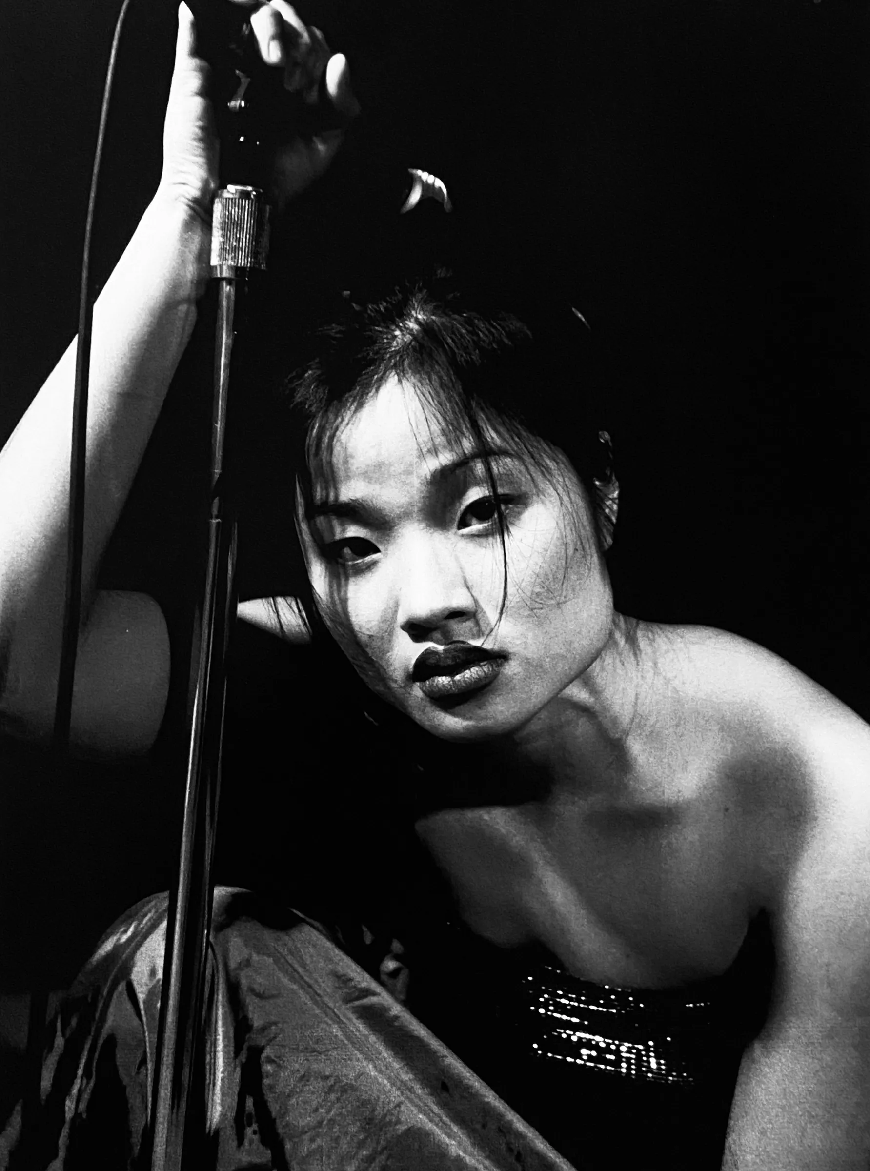 Black-and-white portrait of a woman crouching and gripping a microphone stand, looking directly at the camera with an intense expression, wearing a strapless sequined top.